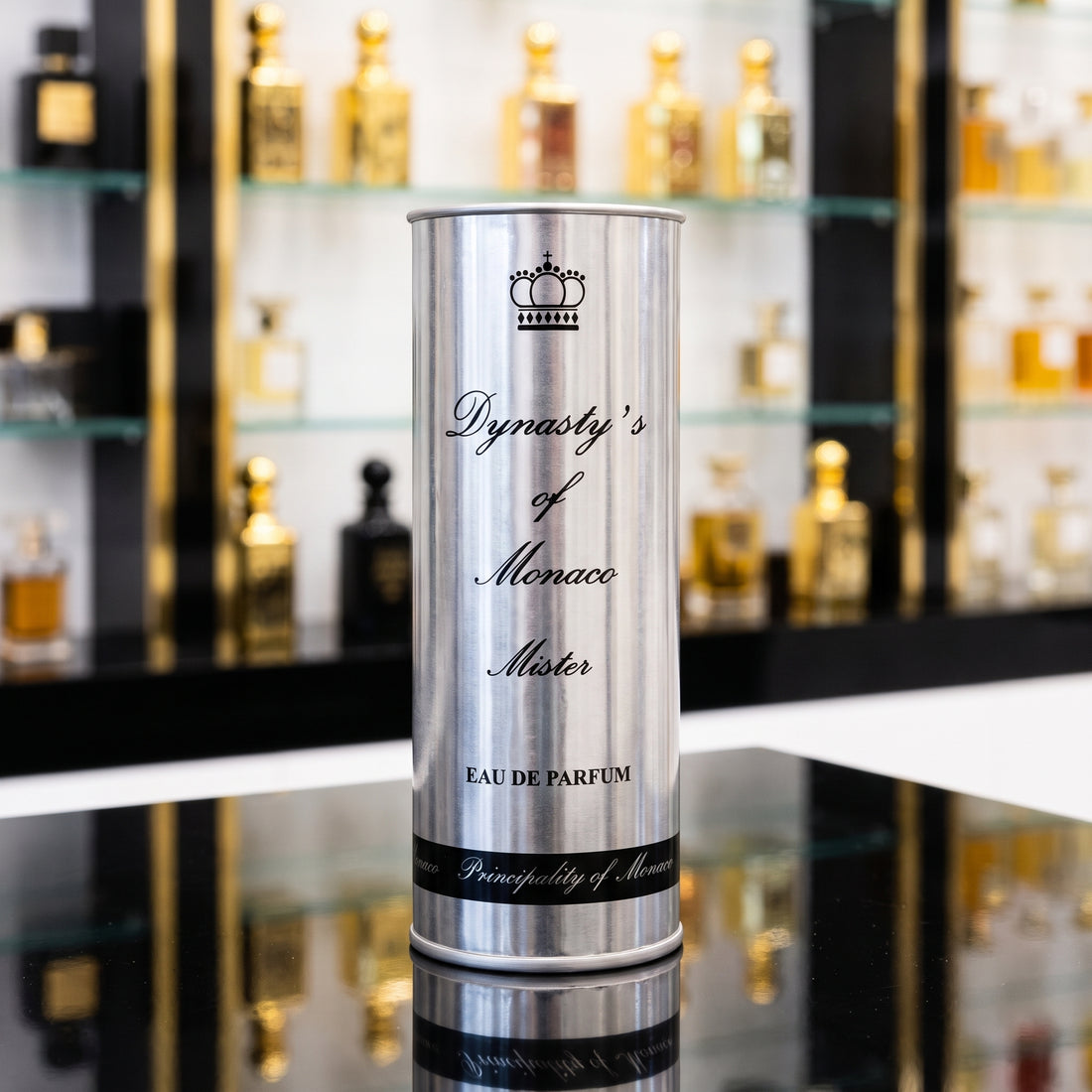 A tall cylindrical silver perfume bottle labeled 'Dynasty's of Monaco Mister' stands on a reflective black surface with a blurred perfume display in the background.