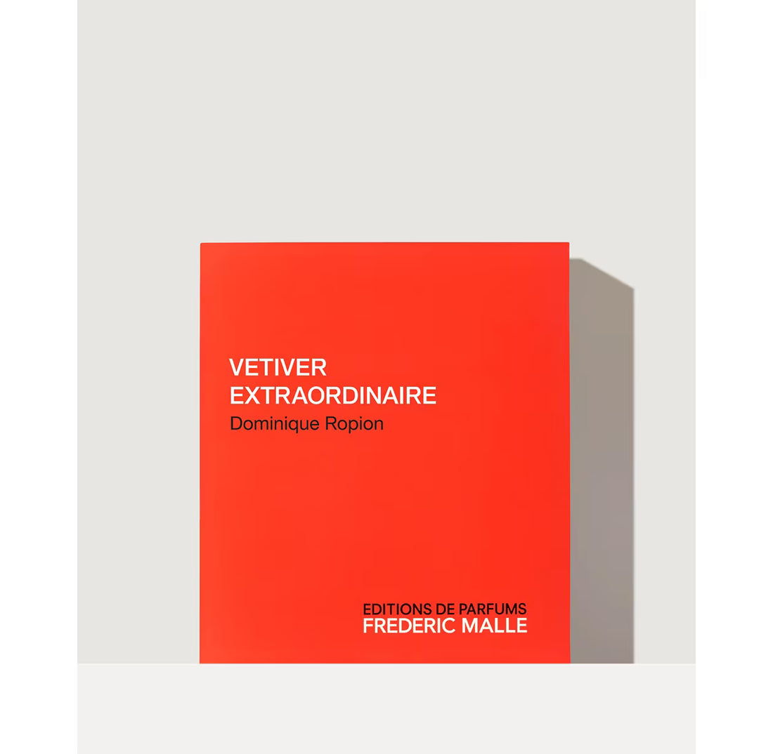 A minimalist perfume box or packaging cover in a vivid orange-red tone, shown standing against a light gray wall with a soft shadow. White text on the front reads “VETIVER EXTRAORDINAIRE,” “Dominique Ropion,” and “EDITIONS DE PARFUMS FREDERIC MALLE.